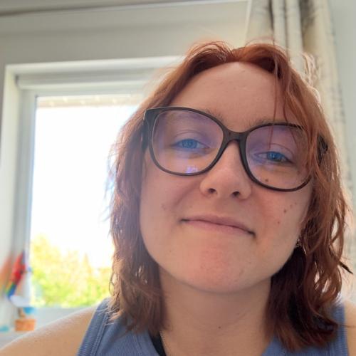 Ellie has shoulder length hair and is wearing brown glasses and a pale blue vest top