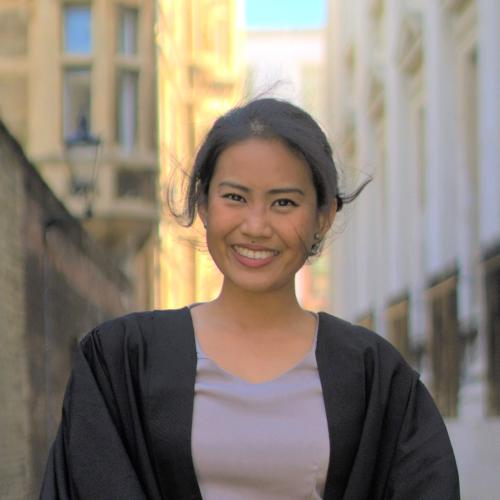 A smiling Marianne stands in a narrow street wearing a black graduation gown.