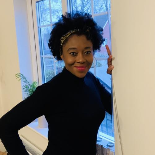 Isang Awah has black hair held back with a hair band, is wearing a black sweater and leading against a window