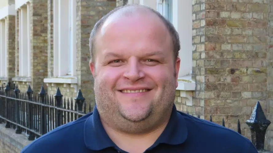 Man with broad smile wearing a dark blue shirt and jumper, and standing in front of brick buildings