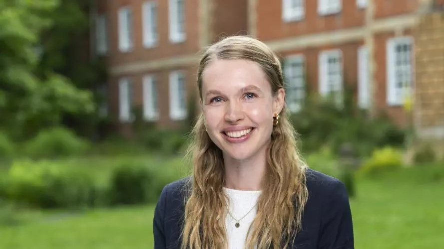 Sophia has long blonde hair. She is wearing a white shirt and navy blazer. She is sat outside in a garden with a red-brick building behind her.