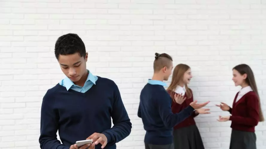 A teenage student stands alone looking down at his mobile. Behind him, three other students stand chatting and laughing