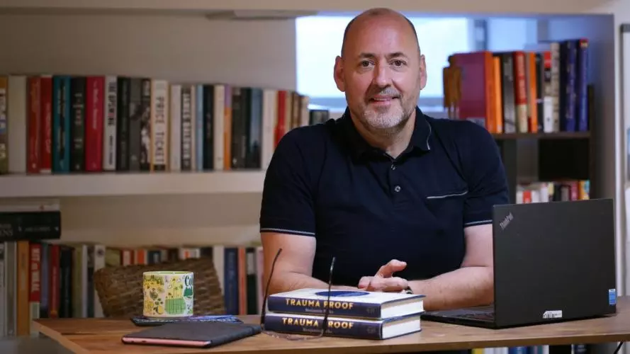 A smiling man seated at a desk in front of bookshelves, wearing a dark short-sleeved polo shirt.