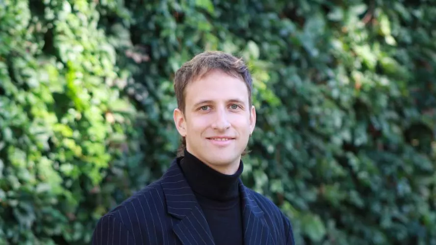 Lorenz Meister has short light brown hair and is wearing a dark turtleneck with navy blue pinstripe blazer. 