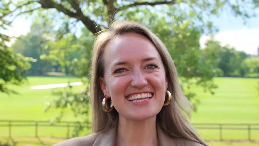 A smiling woman outdoors in a sunlit green park, wearing gold hoop earrings, a taupe blazer, and a pink-and-white patterned top. Trees and an open grassy field are softly blurred in the background.