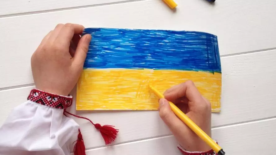 Hands colouring the Ukrainian flag blue and yellow on white paper, with coloured pencils nearby.