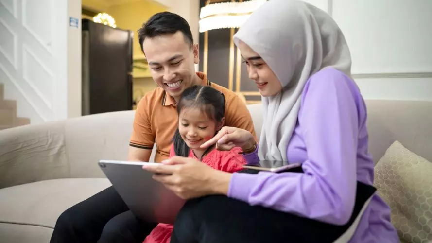 A mother and father relaxing with their young daughter, holding a digital tablet and sitting on a sofa