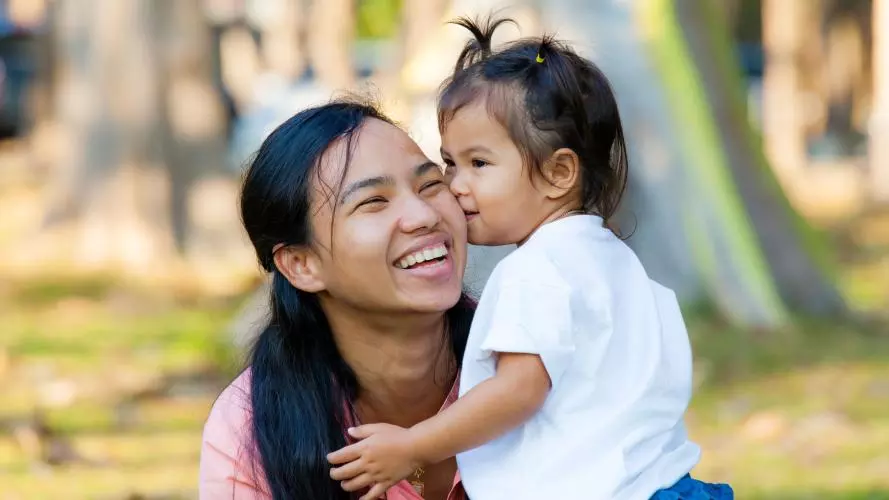A mother crouching down next to her young daughter, who is kissing her check. The mother is laughing. 