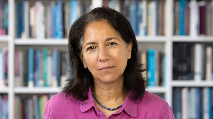 Rebecca Surender has shoulder-length dark hair. She is wearing a pink shirt, sitting in front of some bookshelves