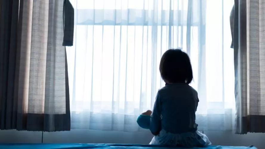 The silhouette of a small child sitting on a bed holding a doll, facing a window with light coming through the curtains.