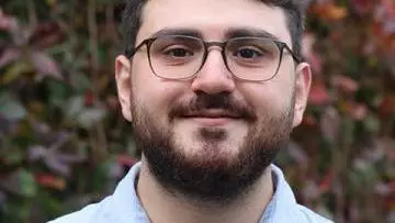 Ertugrul has short brown hair, a brown beard and glasses. He's wearing a pale blue shirt