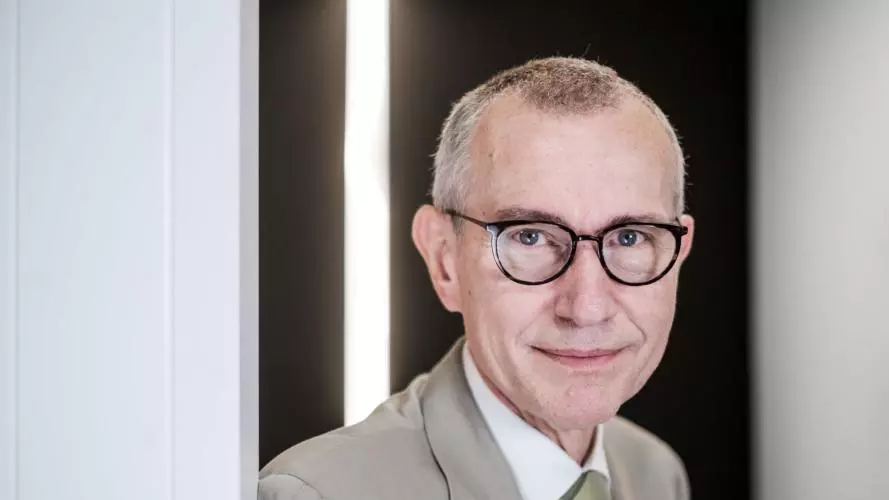 Professor Frank Vandenbroucke is wearing a light suit and round glasses. He is leaning against a light wall.