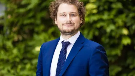 A man wearing a blue suit with brown curly short hair looking at the camera.