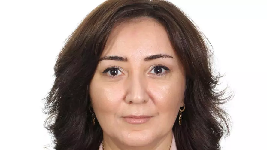 Bahija Aliyeva wears a peach blouse with a floral pattern. She had shoulder-length dark brown hair and is on a plain white background.