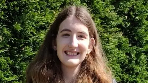Chloe Brimicomb has shoulder-length brown hair and is wearing a blue jacket, standing in front of a green background
