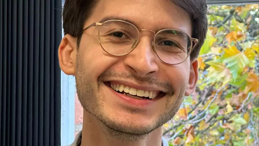David has short dark hair, is wearing glasses and a cream coloured jumper. There is a window behind him with autumnal trees outside.