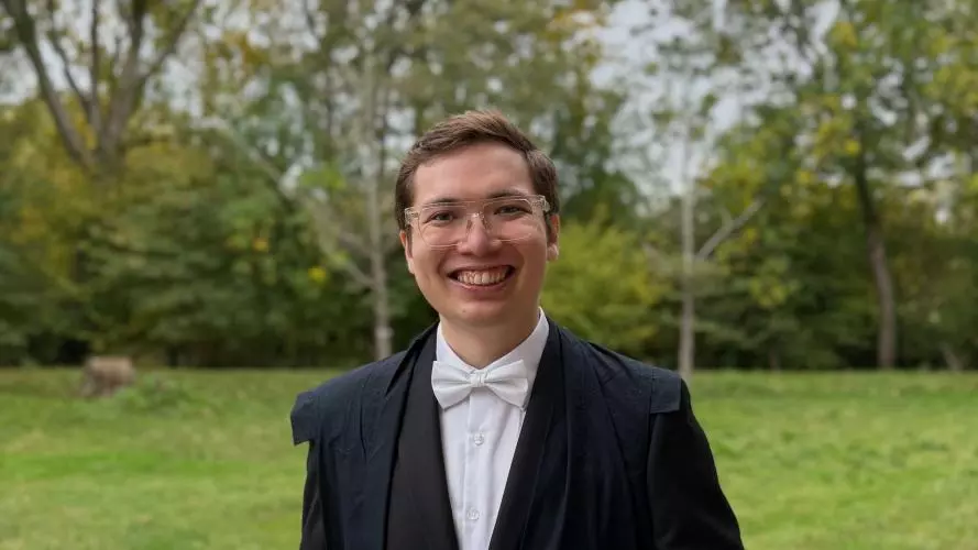 George Rappa has short brown hair and glasses. He's wearing an academic gown, with dark suit and white tie, and is standing in a college garden