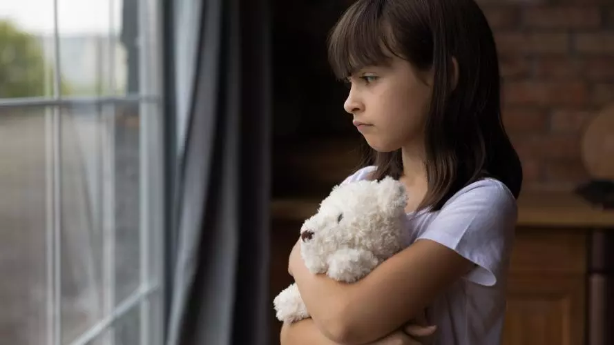 A young girl standing at a window, hugging a teddy bear. Credit: fizkes / Getty Images 1300371821