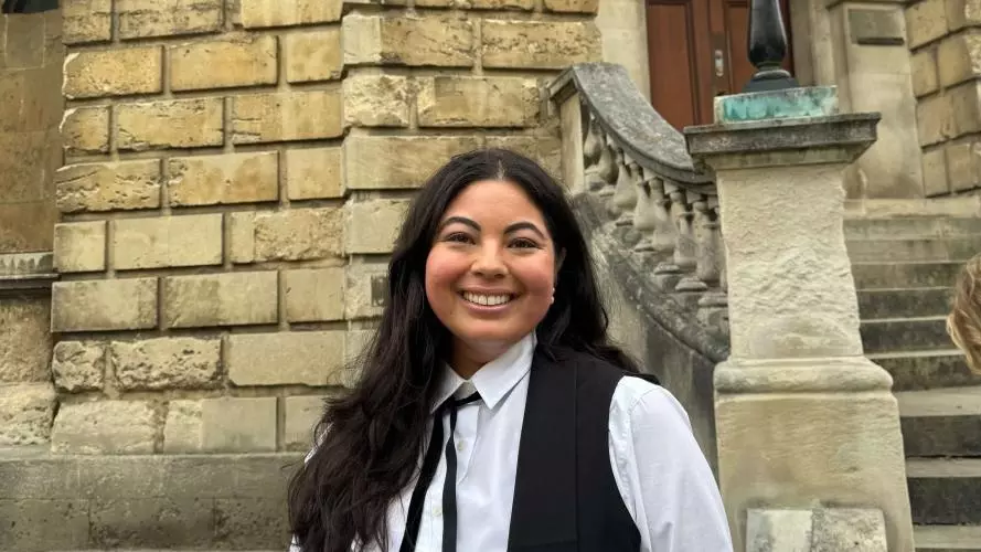 Lhori Webster is wearing academic dress and stands in front of a sandstone building in Oxford. 