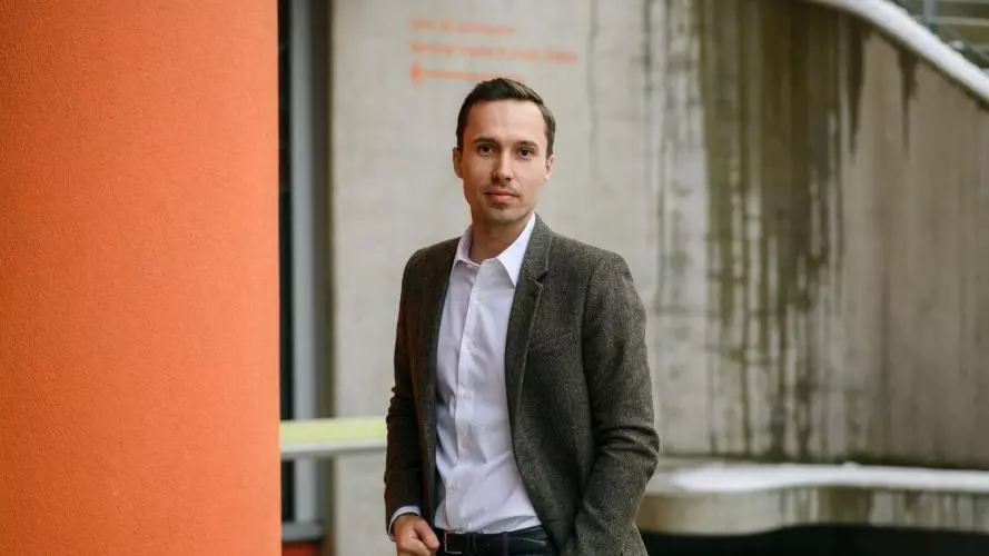 Lukas Lehner wears a white shirt and dark jacket, he stands on an angle next to an orange wall.