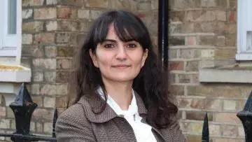 Merve is smiling and standing with her arms folded in front of a brown brick building. She has long dark hair and a fringe. She is wearing a white shirt and brown blazer. 