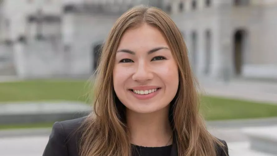 Miya has shoulder-length light brown hair and wearing a dark blazer. She is standing in front of a set of steps