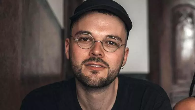 steffen has short black hair, black beard and metal-rimmed glasses. He's wearing a black jumper 