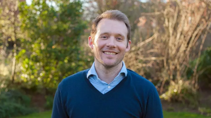 Alex Sutherland has short fair hair and is wearing a light blue shirt with a navy jumper. He is standing outside in a garden, smiling at the camera. 