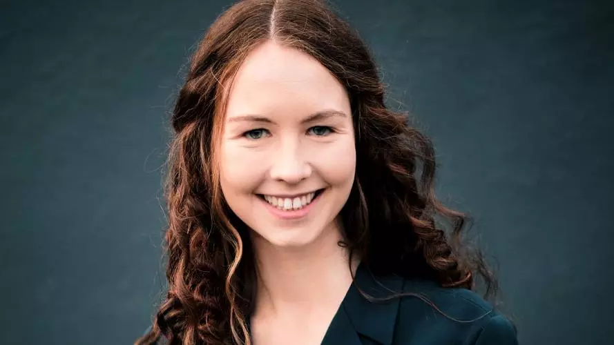 Tamsin Greene Barker is smiling at the camera, with long, dark, shoulder-length curly hair, wearing a dark jacket.