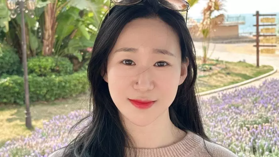 Lin Chen has long dark hair. She is standing in front of some flower beds and foliage and is wearing a beige knitted jumper. 