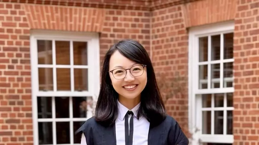 Yiwei Zhou stands in a courtyard wearing academic dress. She is smiling, holding a mortar board.