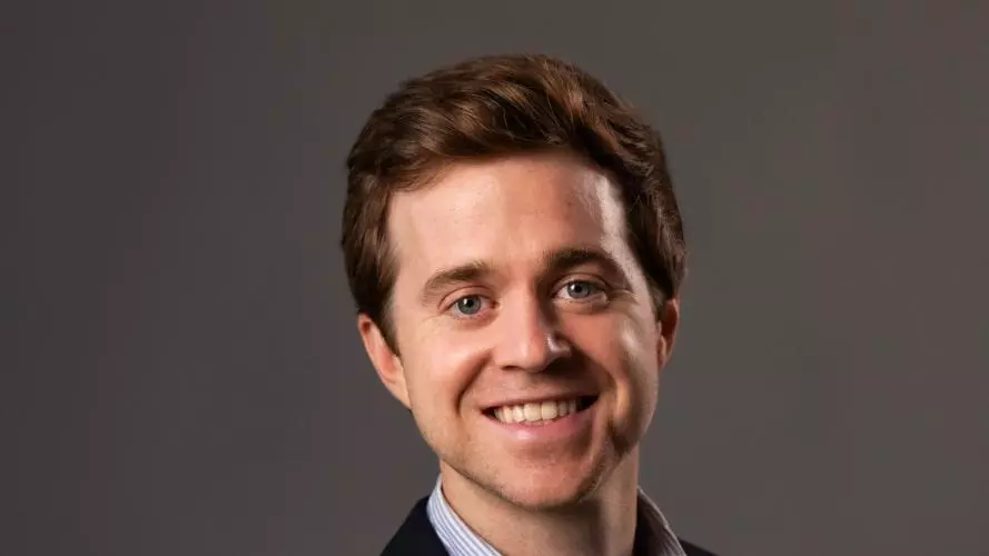 Zach Parolin has short brown hair. He is wearing a blue shirt and dark blazer, standing against a dark background. He is facing the camera and smiling.
