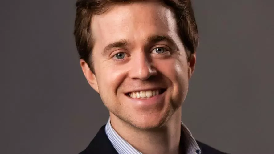 Zach Parolin has short brown hair. He is wearing a light blue shirt and a dark blazer, and smiling broadly. 
