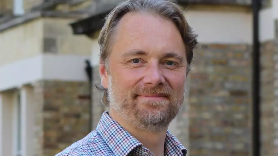 Mark Fransham is wearing a plaid shirt, with short blonde hair, a short bear and mostashe and smiling gentle at the camera.