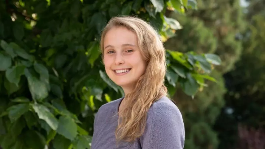 Charlotte Evans is wearing a purple top , standing sideways at the camera, with shoulder length blonde hair smiling at the camera.