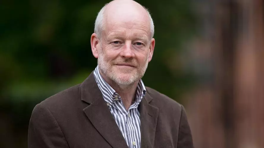 Brian has short grey hair and beard. He's wearing a white and purple stripped shirt with dark jacket 
