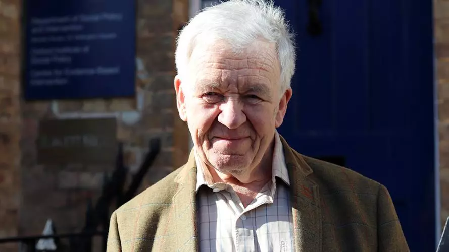 David has grey hair, and he's wearing a white checked shirt with light brown jacket