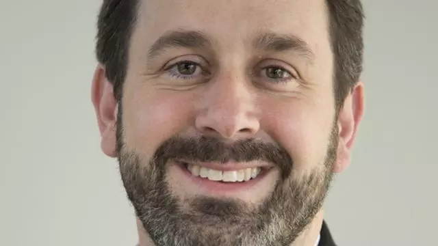 David has short dark hair and a beard, he is smiling at the camera, and wearing a white checked shirt with a dark jacket. 