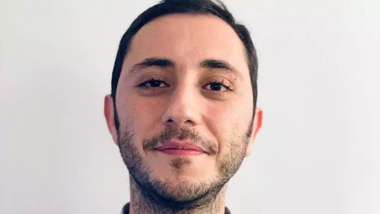 Dr Bedük has short dark hair. He is wearing a light blue shirt and grey jumper. 