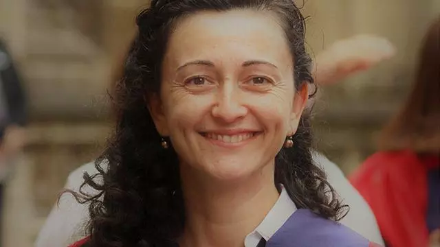 Elona  has long dark curly hair and is smiling at the camera. She's wearing a blouse under a purple and red jacket