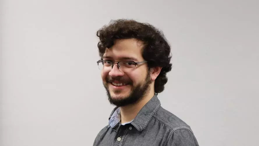 Man with curly dark hair and beard and wearing dark rimmed glasses and grey shirt