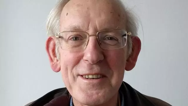 Smiling man with short grey hair, bald head wearing a dark jacket and with gold rimmed glasses.