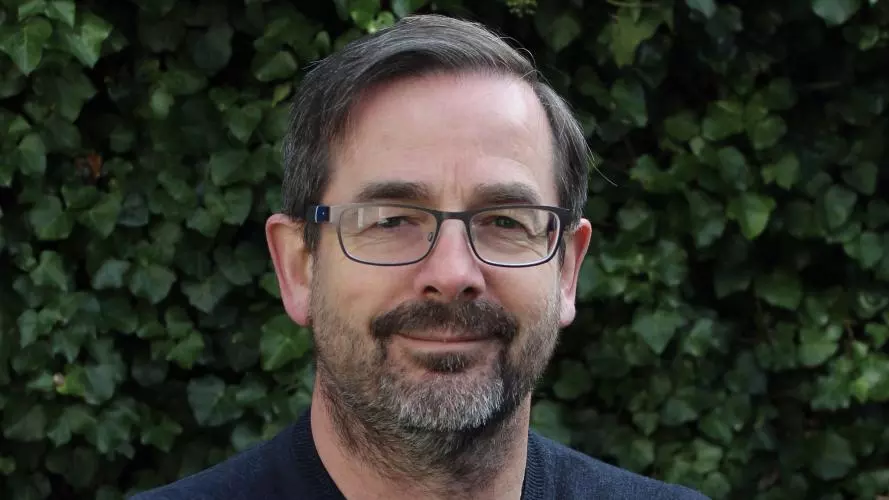 Gerry has short dark hair and a beard. He's wearing glasses and a navy blue jumper. He's standing in front of a hedge. 