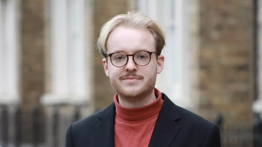 Hendrik Schäfer has blonde hair and wears glasses. He is standing in front of the DSPI building in a red polo-neck top and blazer.