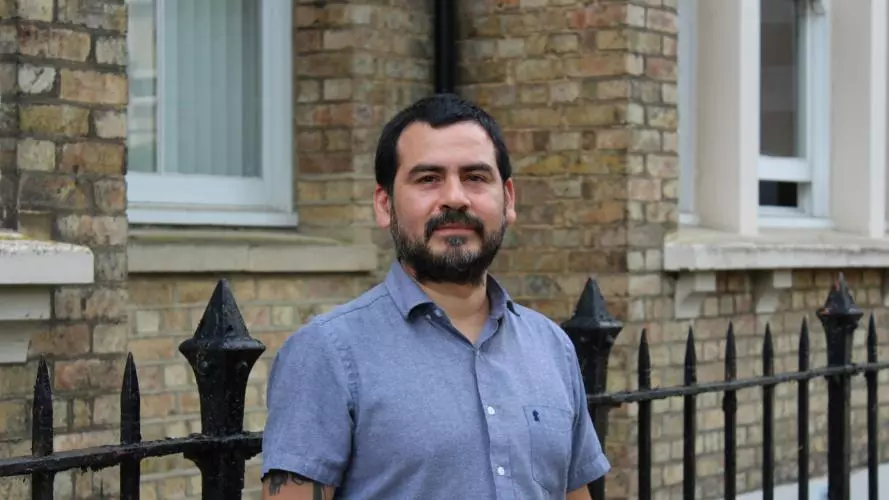 Dr Munoz has short black hair and black facial hair. He is wearing a short-sleeved blue shirt and standing outside a brick building. 