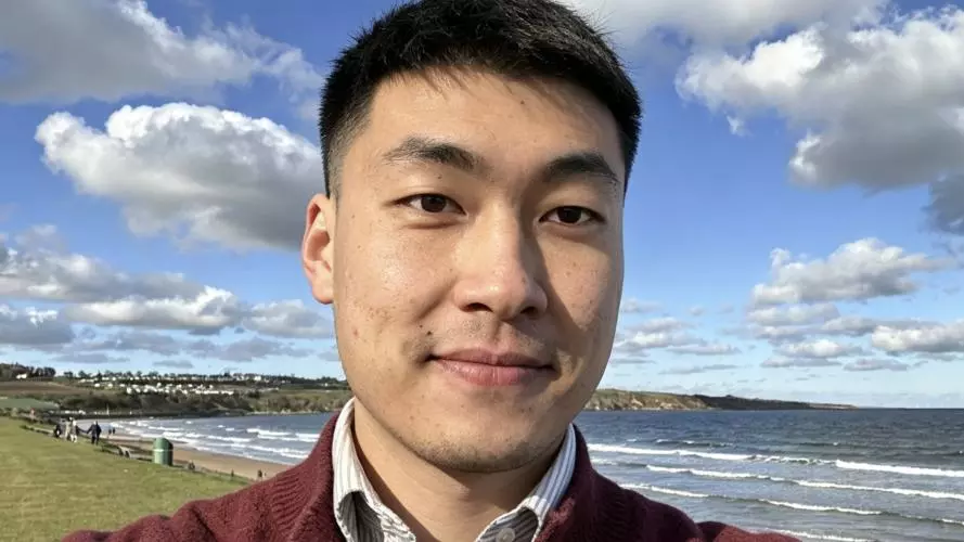 Man with short dark hair, wearing a jumper and standing near the sea