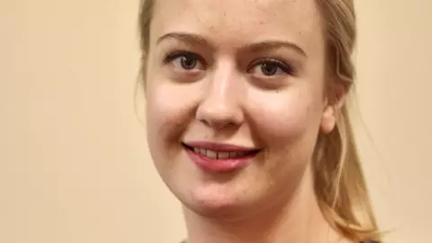 katariina has long blonde hair tied back in a ponytail. She's wearing a buttoned-up navy blue cardigan