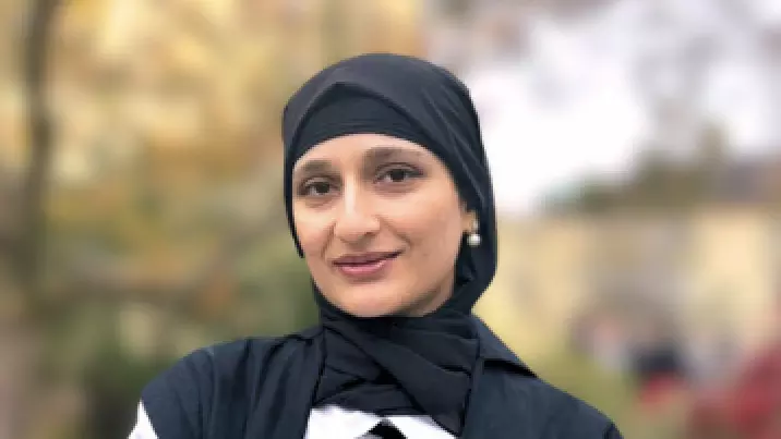 Kayanat Iltaf stands with folded arms wearing academic dress in front of an out-of-focus tree in the background. 