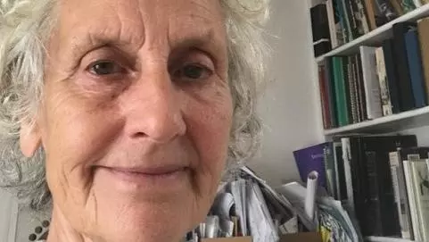 Dr Peretz has short curly white hair. She is sat in front of a large bookcase, smiling. 