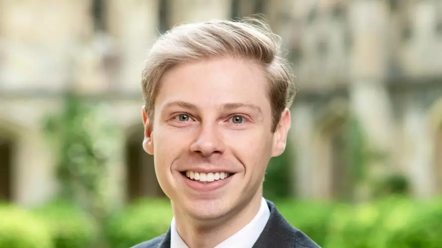 Madison Little has short blonde hair. He is wearing a white shirt, dark tie and blazer. He is standing in front of some grass in a courtyard.